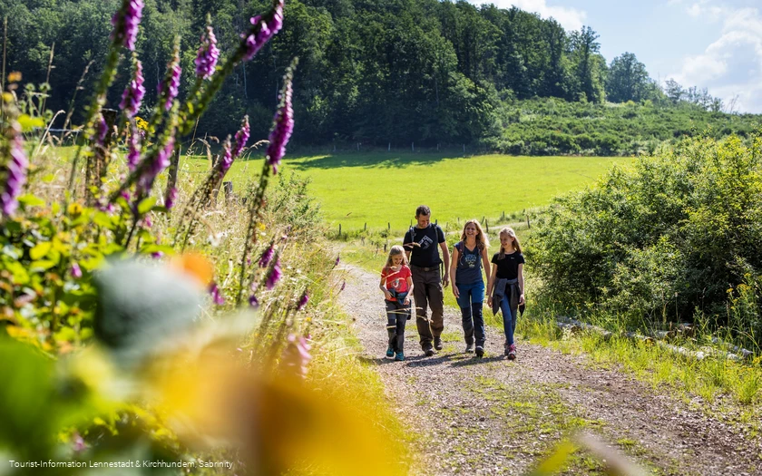 Familie wandert auf Wanderweg zwischen bunt blühenden Blumen und Wiesen.